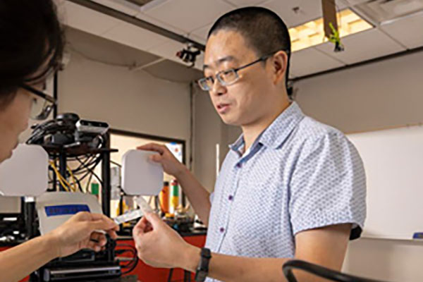 professor jian zhang in glasses and a short-sleeved shirt discusses a small handheld device with a woman in a lab, conveying focus and collaboration.