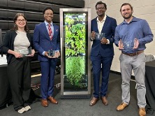 Four people stand smiling with awards in front of a vertical hydroponic garden. They are dressed formally, expressing a celebratory and professional tone.