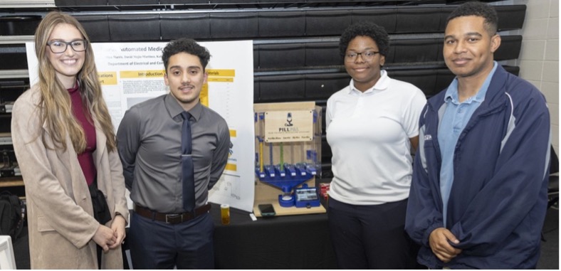 Four people stand smiling in front of a science project display featuring a poster and a wooden model with tubes labeled "P.U.L.S.E." in a classroom.