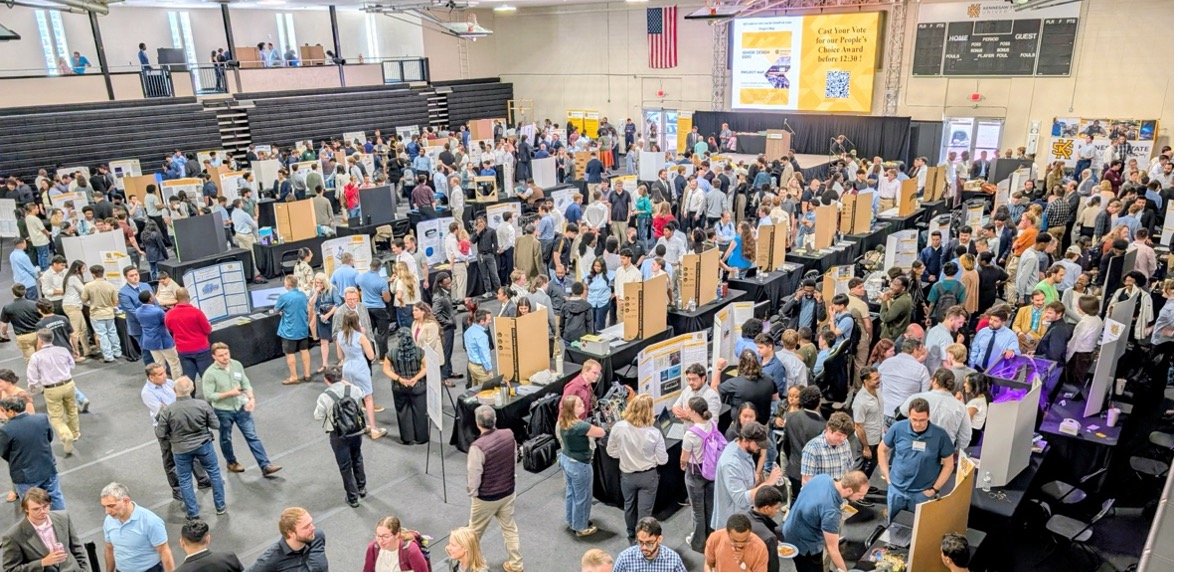A bustling exhibition hall with numerous attendees browsing booths displaying posters and projects. The atmosphere is lively and collaborative, with people engaging in discussions. An American flag hangs on the wall, and a large banner features prominently in the background.