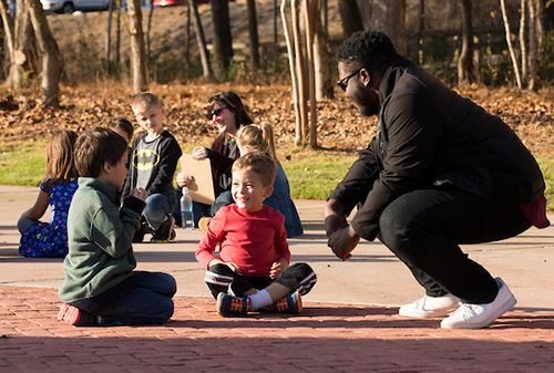 A man is kneeling on the ground, surrounded by children who are attentively listening and participating.