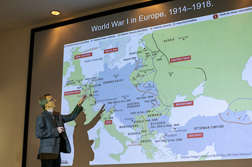 A professor stands confidently in front of a large projection screen, engaged in a presentation or discussion.