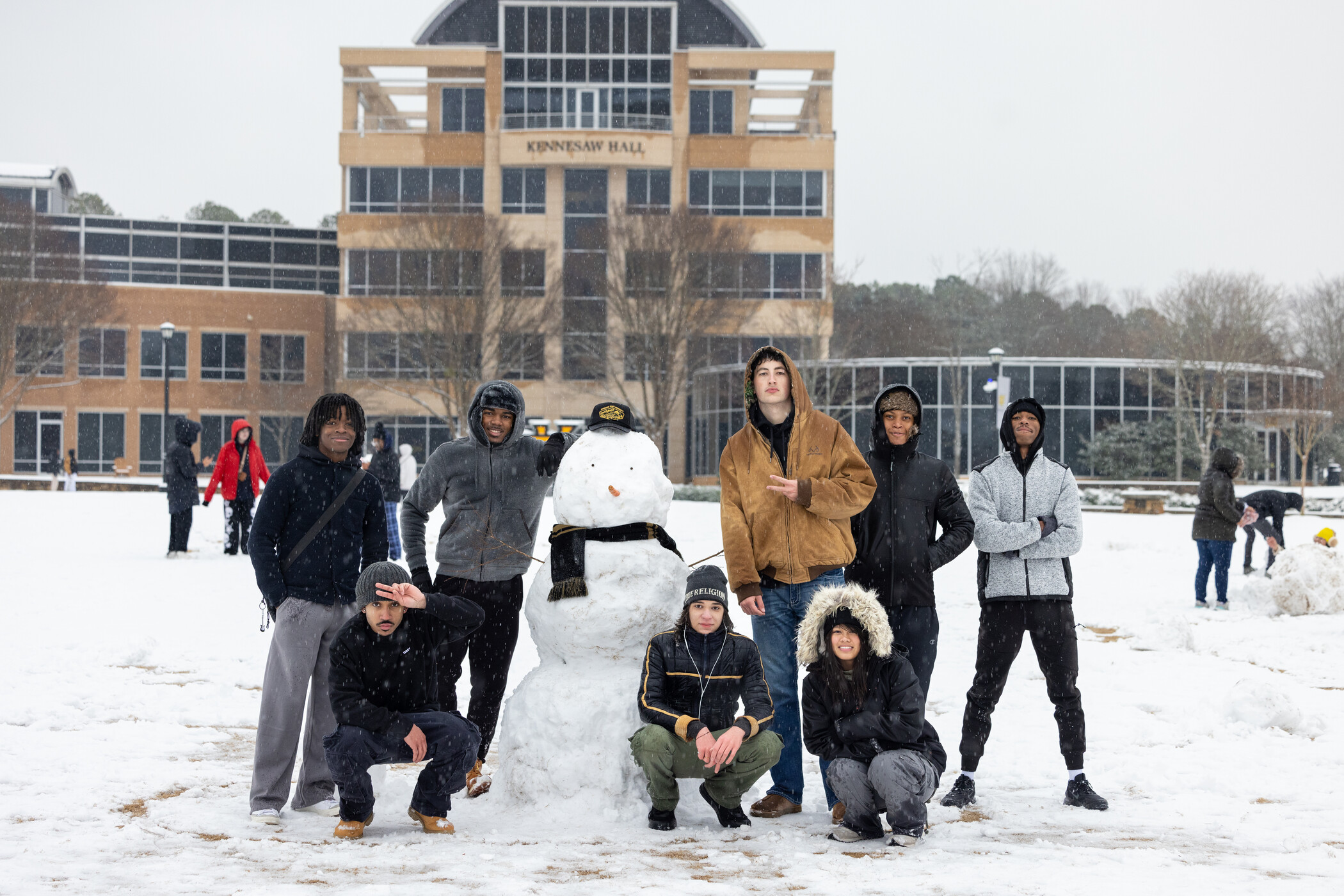 A group of KSU students meet on the green to pose with a snowman wearing KSU gear on a campus snow day.