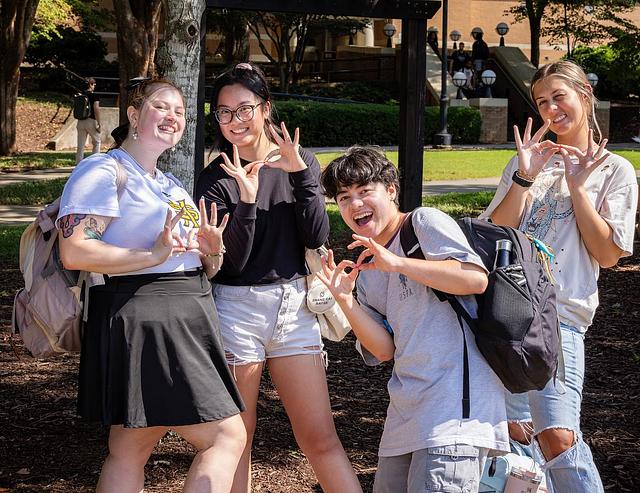 A group of four students outside on the campus green doing the "owl eyes" symbol with their hands