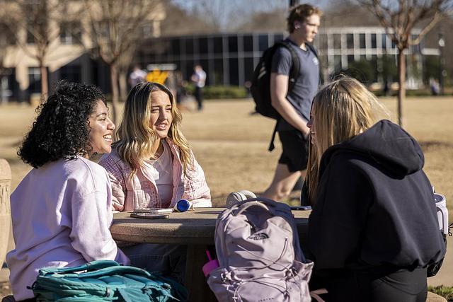 A group of three students sitting and talking at an outdoor table by the campus green