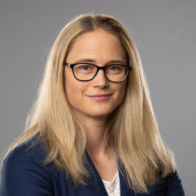 Photo of a woman with long blonde hair and black glasses, wearing a business coat