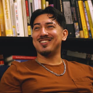 Photo of a smiling Hispanic male sitting casually in front of a library bookshelf