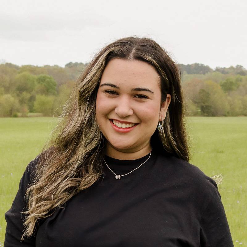 Person with long wavy hair wearing a black top stands outdoors in a grassy field with trees in the background.
