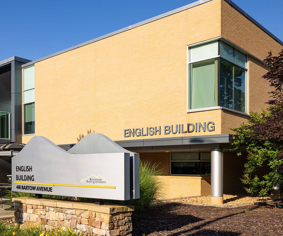 Exterior view of the English Building at Kennesaw State University, featuring a modern two-story structure with tan brick walls and large glass windows. A gray sign in front reads ‘English Building, 440 Bartow Avenue’ with the Kennesaw State University logo, mounted on a stone base surrounded by landscaping.