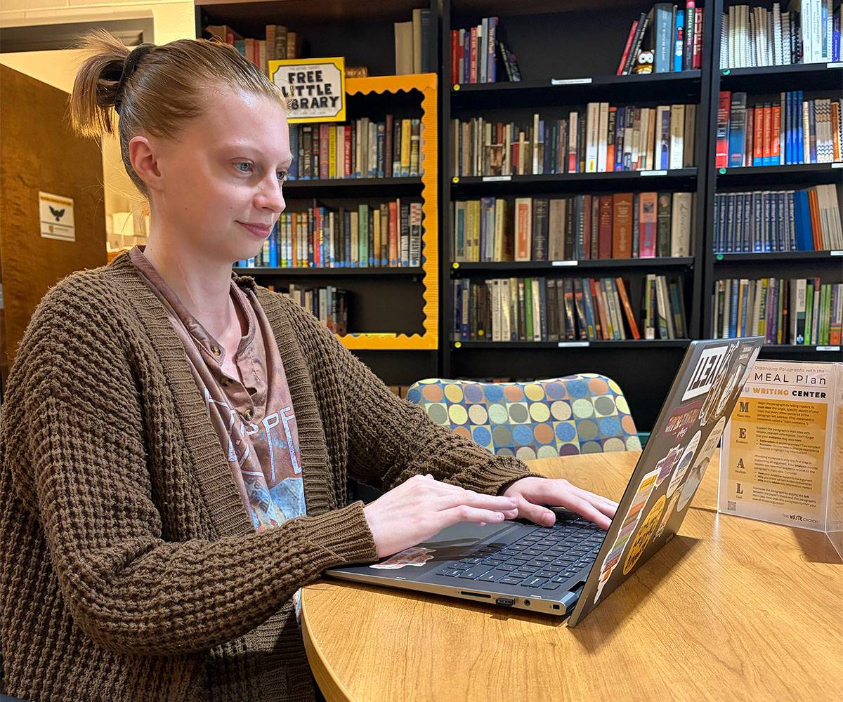 Writing assistant at KSU Writing Center typing on a laptop during an online session, with bookshelves in the background.
