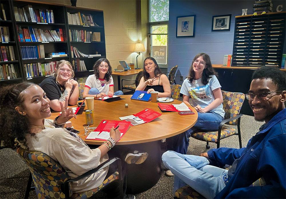 Students gather around a round table in the Writing Center, engaged in discussion and working with notebooks, folders, and drinks.
