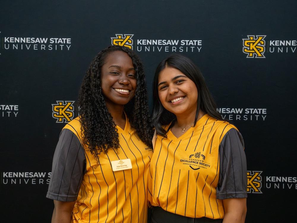  / Two Kennesaw Excellence Society members pose in front of a KSU-branded wall. 
