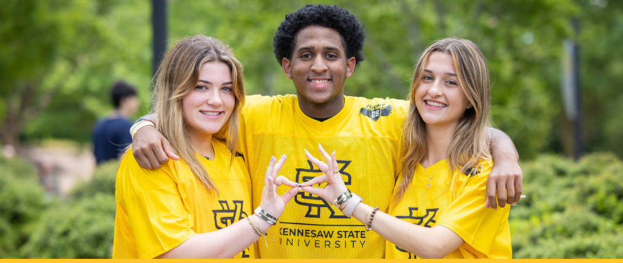 Three students in yellow Kennesaw State University shirts stand outdoors, arms around each other, smiling.