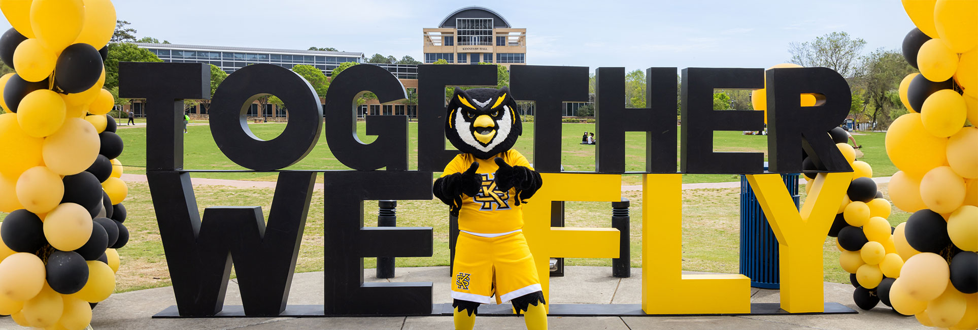 KSU mascot Scrappy the Owl in a gold and black uniform stands in front of large block letters spelling "TOGETHER WE FLY" on a grassy campus area.