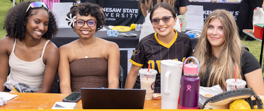 Four smiling KSU students sit behind a table at an outdoor Kennesaw State University event, promoting club sports.
