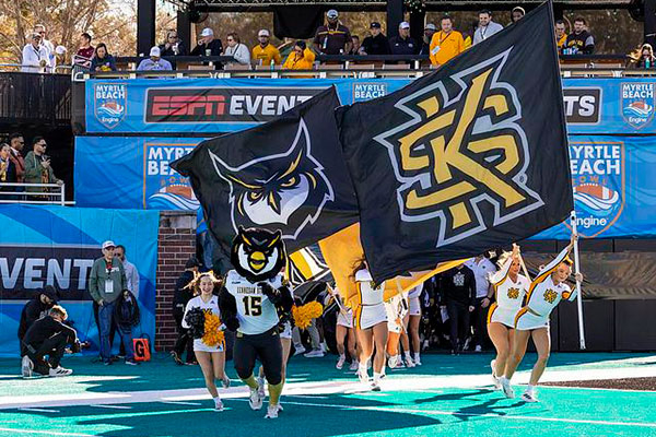 Cheerleaders and a ksu mascot run onto a football field. They wave large black flags with yellow and gold logos, under ESPN and Myrtle Beach event banners.