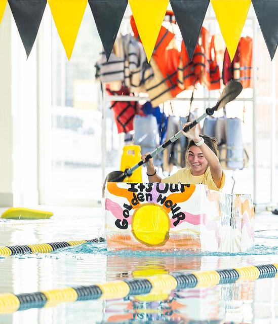 student in cardboard boat paddling in pool race