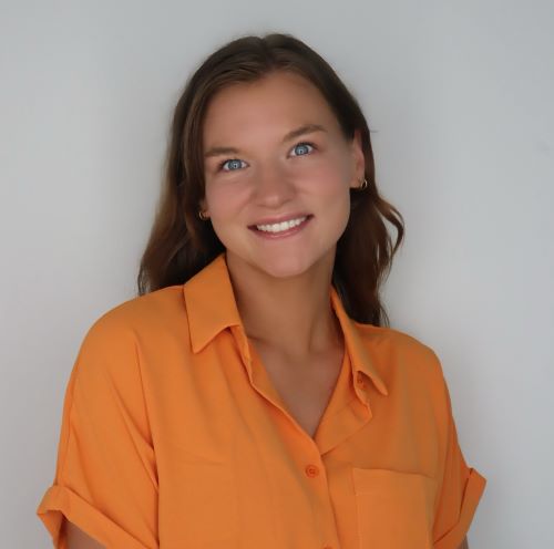 A headshot image of DJ. A person with long wavy brown hair is shown from the shoulders
                                    up, wearing a bright orange short-sleeve button-up shirt against a plain light background.