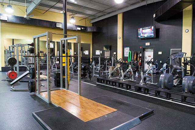 Spacious gym with various exercise equipment, including weights, treadmills, and bikes. Bright lighting and a TV create an energetic atmosphere.