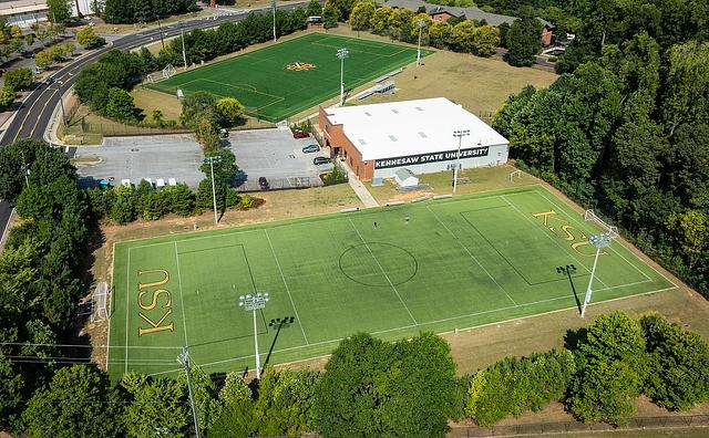 Aerial view of Kennesaw State University sports fields. Two green fields labeled "KSU" are surrounded by trees and a road, near a white building.