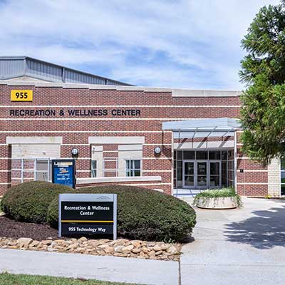 Recreation and Wellness Center entrance with red brick facade, large glass doors, and sign displaying the address 955 Technology Way under a blue sky.