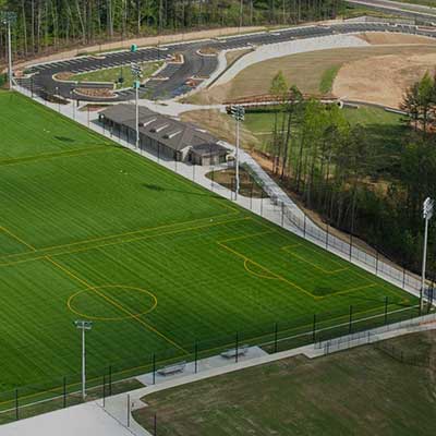 Aerial view of an outdoor sports complex featuring a vibrant green soccer field with yellow lines. Surrounding are parking lots and wooded areas.