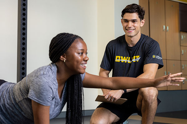 Woman in a gray shirt performs a stretching exercise on the floor, smiling. A man in a black "OWLFIT" shirt kneels beside her, holding a clipboard, also smiling.