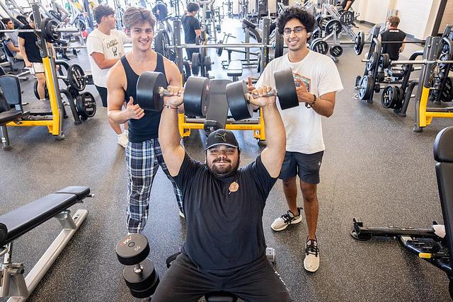 Three men in a gym, one seated and lifting dumbbells while two others stand beside him, smiling. The gym is busy with equipment and people exercising in the background. The mood is energetic and motivating.