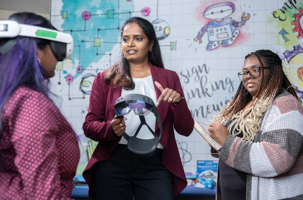 two students with talking to a professor, one student is taking notes and the other is wearing a virtual reality headset