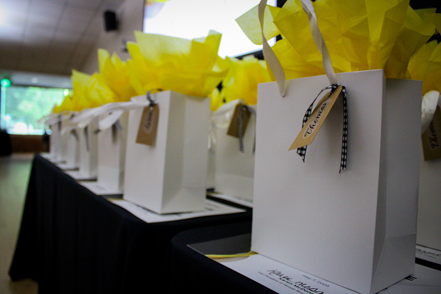 HILP Dinner 2025-2026 / A line of white gift bags with yellow tissue paper and name tags, adorned with black-and-white checkered ribbon, placed on a table, creating a festive atmosphere.