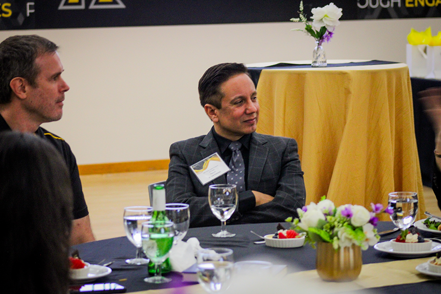 HILP Dinner 2025-2026 / A group of people in formal attire sit at a round table set with drinks and desserts. A man in a checkered suit appears engaged, with flowers on the table.