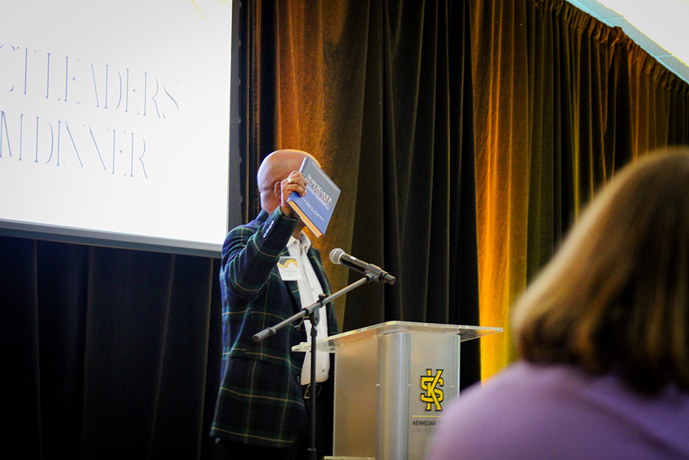 HILP Dinner 2025-2026 / A speaker in a plaid jacket holds up a book at a podium with Kennesaw State University branding. A large screen and audience are visible.