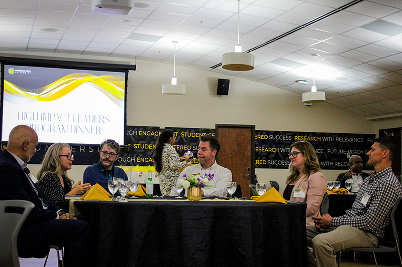 HILP Dinner 2025-2026 / A group of people sit around a table with black tablecloth and yellow napkins, engaged in conversation at a "High Impact Leaders Program Dinner." A large screen displays the event title in the background. The setting is formal, with a modern, well-lit room, creating a professional and convivial atmosphere.