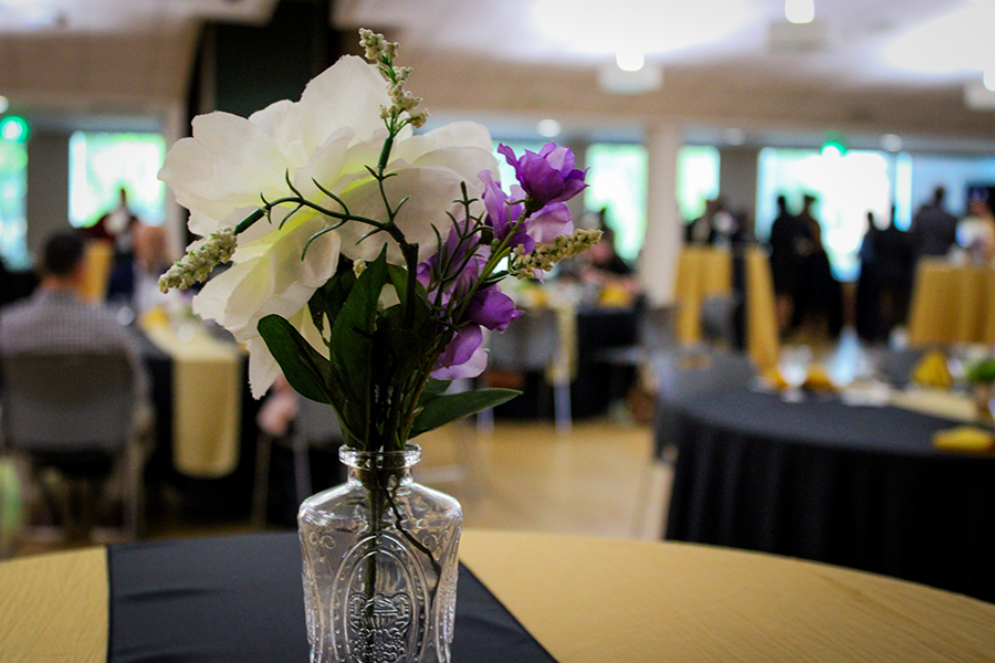 HILP Dinner 2025-2026 / A vibrant conference room with tables and chairs filled with people engaged in conversation. A vase with purple and white flowers is in the foreground.