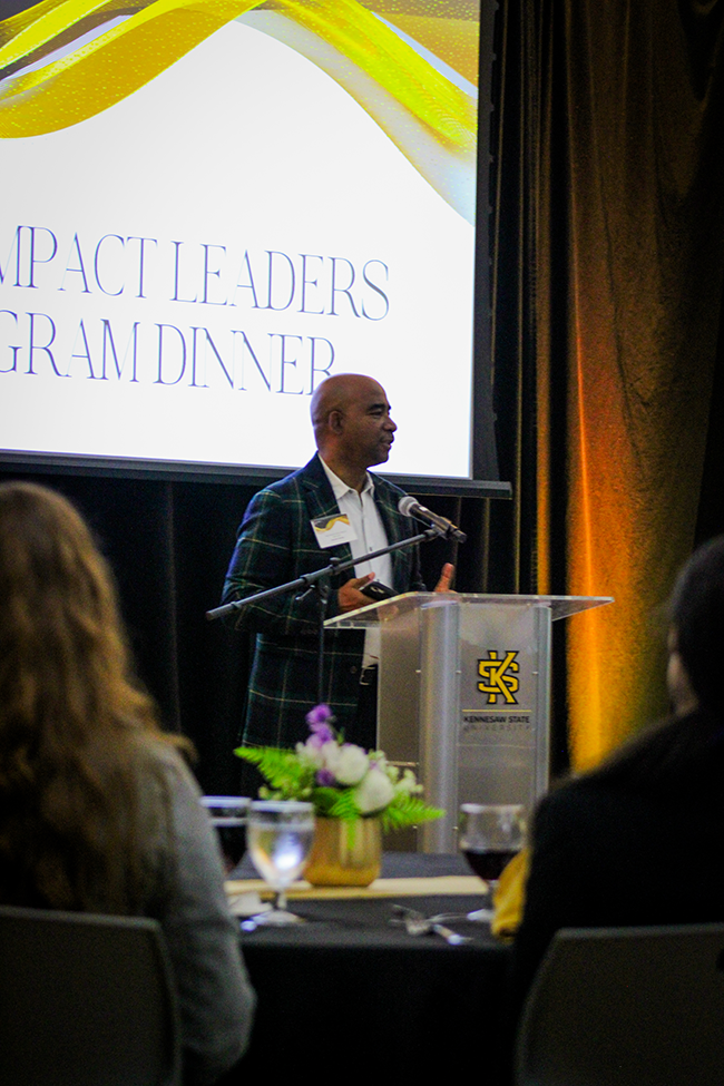 HILP Dinner 2025-2026 / A man speaks at a podium with a microphone, in front of a screen reading "High Impact Leaders Program Dinner." Attendees watch, with flowers in view.