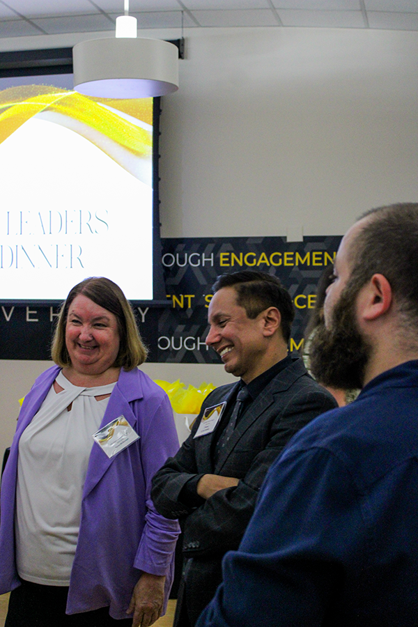 HILP Dinner 2025-2026 / Group of three people smiling and chatting at a networking event. Background shows a large screen with "High Impact Leaders Program Dinner." Jovial atmosphere.