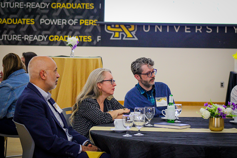 HILP Dinner 2025-2026 / Three people sit at a round table with glasses and a book, attentively listening. Banners read “Future-Ready Graduates.” The atmosphere is professional.