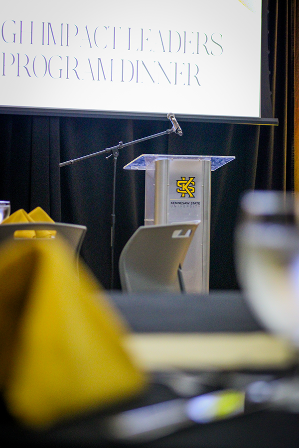 HILP Dinner 2025-2026 / A podium with a microphone stands in front of a large screen displaying "High Impact Leaders Program Dinner." Tables with yellow napkins are partially visible.