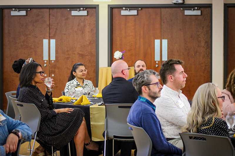 HILP Dinner 2025-2026 / A diverse group of people seated around a table with yellow napkins, engaged in attentive listening during a formal event, conveying a professional atmosphere.