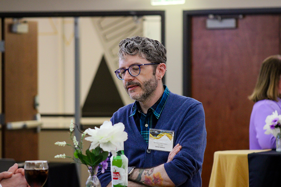 HILP Dinner 2025-2026 / A man with glasses and tattoos sits at a table with a flower vase, engaged in conversation. The setting is casual and social, with a relaxed mood.