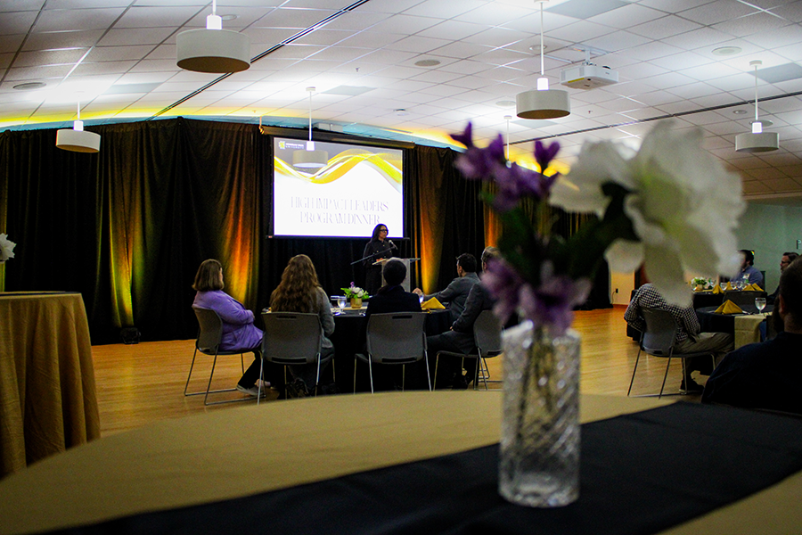 HILP Dinner 2025-2026 / A formal dinner event with guests seated at round tables in a well-lit room. A screen displays “High Impact Leaders Program Dinner,” creating a sophisticated atmosphere.