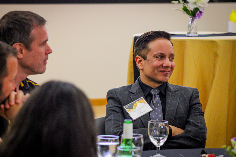 HILP Dinner 2025-2026 / A smiling person in a suit sits at a table during an event, surrounded by glasses and bottles. A floral centerpiece is on a table in the background.