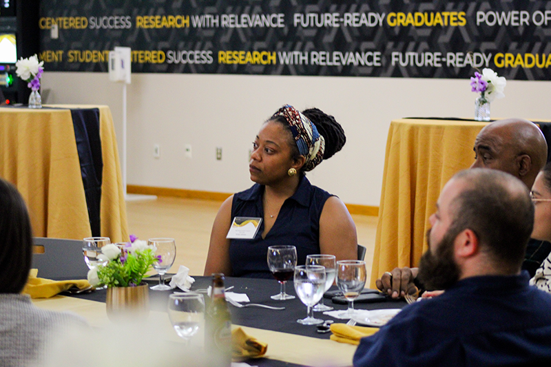 HILP Dinner 2025-2026 / A woman sits at a table during a formal event, attentively listening. The room has motivational banners and elegant table settings, creating a focused, professional atmosphere.
