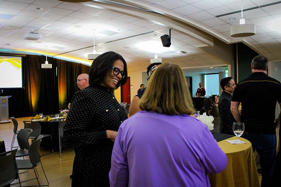 HILP Dinner 2025-2026 / A woman in a polka dot dress smiles while talking to another in a purple sweater at a lively event. The room is well-lit with tables and decorations.