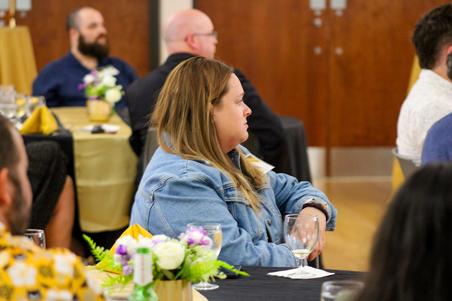 HILP Dinner 2025-2026 / A woman in a denim jacket attentively listens at a formal event. Tables feature black and gold linens, wine glasses, and pastel flower centerpieces.