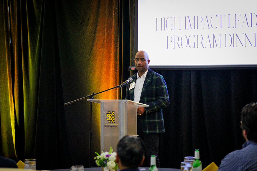 HILP Dinner 2025-2026 / A man in a plaid jacket gestures from a podium with a microphone, against a backdrop with text. The atmosphere is formal and engaging.