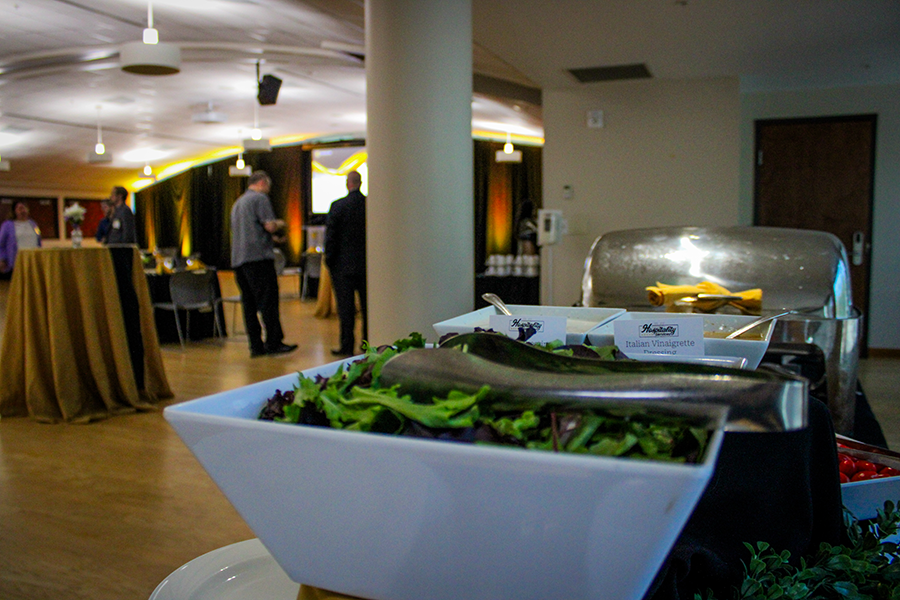 HILP Dinner 2025-2026 / A buffet with a white salad bowl containing leafy greens in the foreground. In the background, a dimly lit room with a few people gathered, creating a relaxed atmosphere.