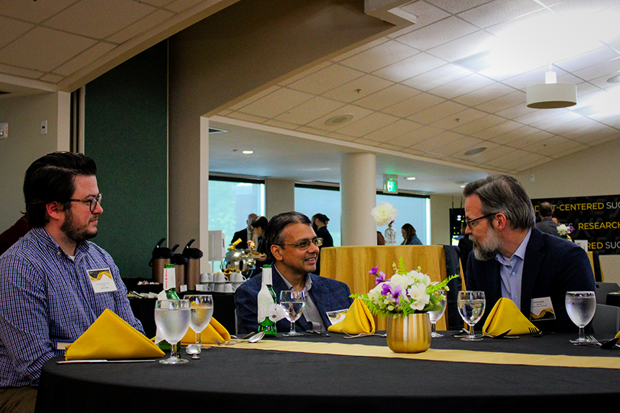 HILP Dinner 2025-2026 / Three men in business attire converse at a table set with yellow napkins and floral centerpiece. The setting is a formal indoor event.