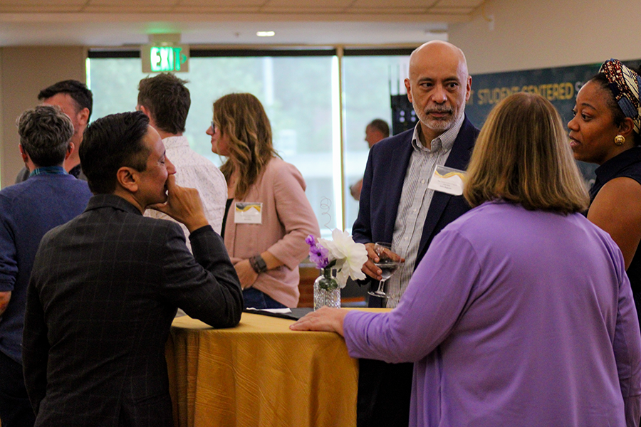 HILP Dinner 2025-2026 / A group of people engage in conversation around a table covered with a yellow cloth at a networking event. They appear attentive and engaged.

