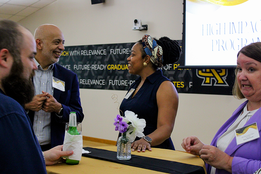 HILP Dinner 2025-2026 / A group of diverse people engages in lively conversation around a table adorned with purple and white flowers in a vase. Background banners read "Future-Ready Graduates."